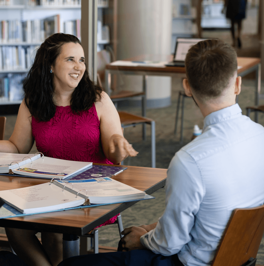 Smiling women and men engaged in a discussion over books on a table in a library.