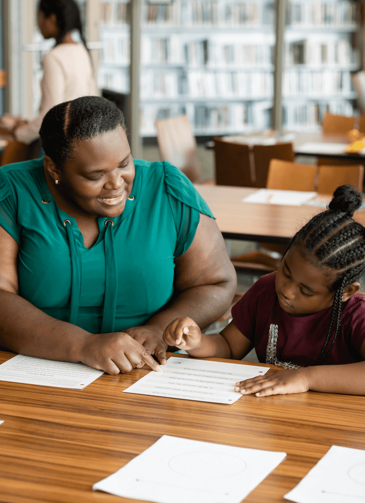 A female teacher instructs a young girl in reading at a classroom desk.