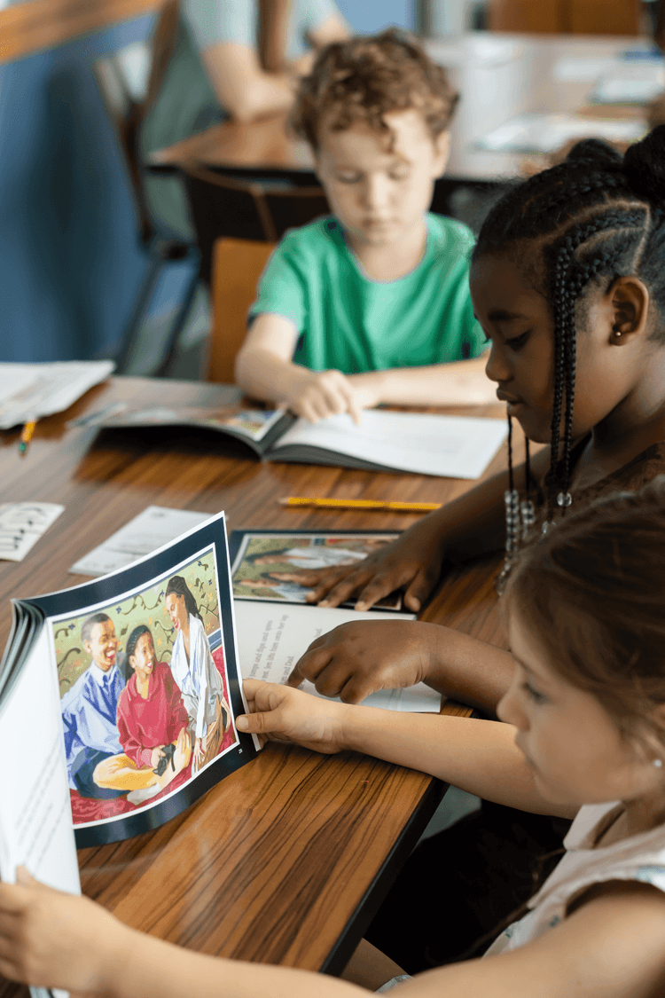 Two young girls and a boy looking at picture books at a classroom desk.