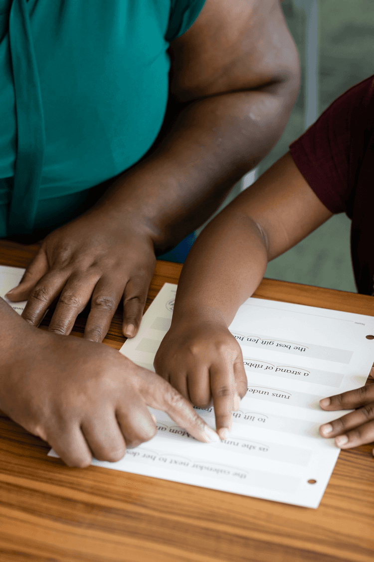 A teacher's hand pointing to a textbook to assist a young girl in the reading process.